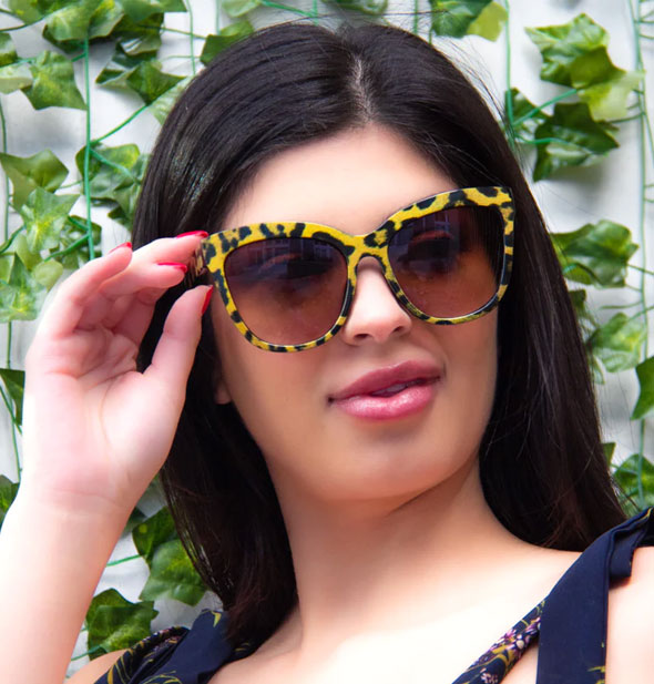 Model wears a pair of gold animal print sunglasses with a cat-eye shape against a backdrop of ivy
