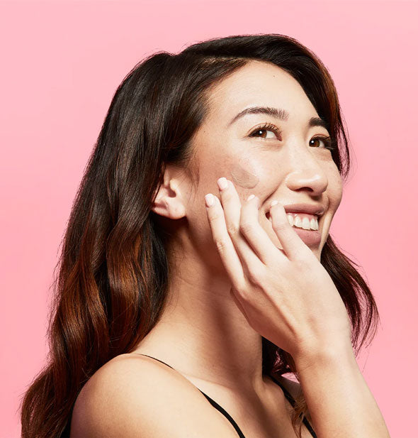Smiling model applies a dab of tinted sunscreen to cheek with fingertip