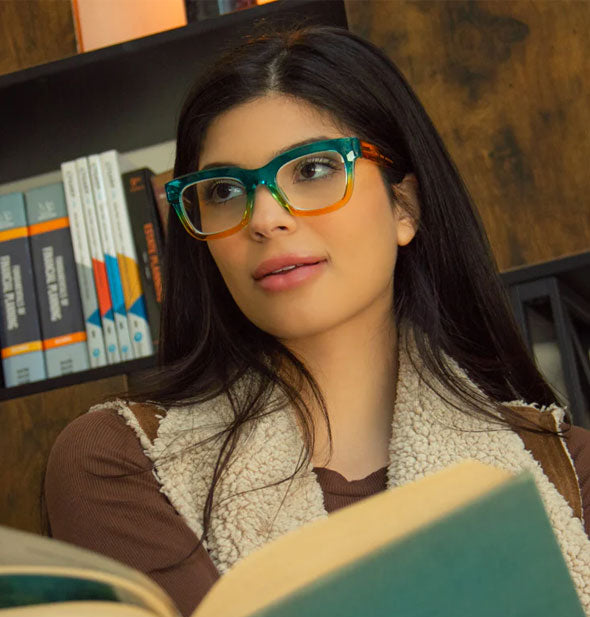 Model holding book wears a pair of thick-rimmed teal and orange ombre reading glasses