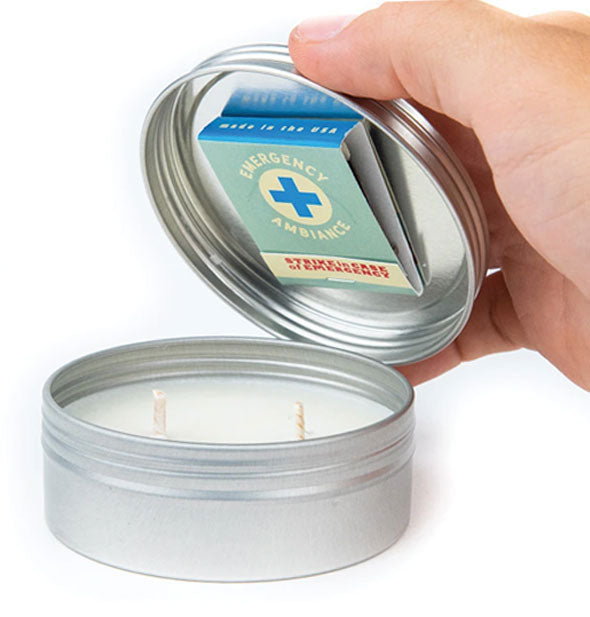 Model's hand lifts the lid of an Emergency Ambiance Travel Tin Candle to reveal a pack of matches attached to its underside