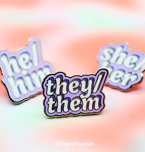 Grouping of three purple enamel pins that say He/Him, They/Them, and She/Her respectively are labeled with Pages Peaches copyright