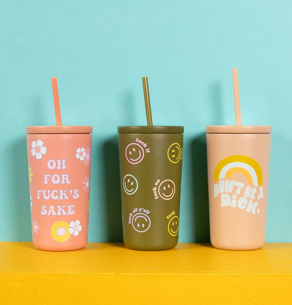 Tumblers with straws in three designs: pink Oh For Fuck's Sake with daisies, olive green "Fuck It" Smileys, and peach rainbow Don't Be a Dick, all on a gold platform against a blue backdrop