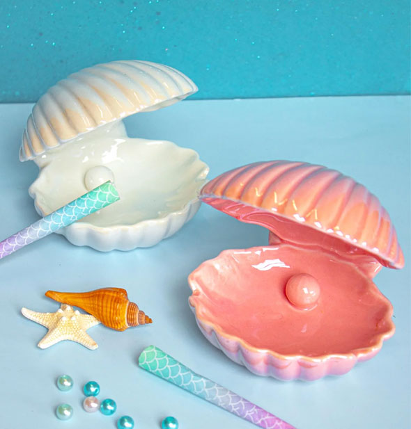 White and pink iridescent ceramic ashtrays resemble open clamshells and are staged on a blue backdrop with seashells, blue pearls, and ombre fish scale cigarettes