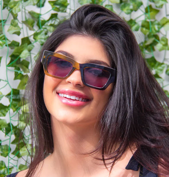 Smiling model wears a pair of half brown, half black square sunglasses in front of an ivy-covered backdrop