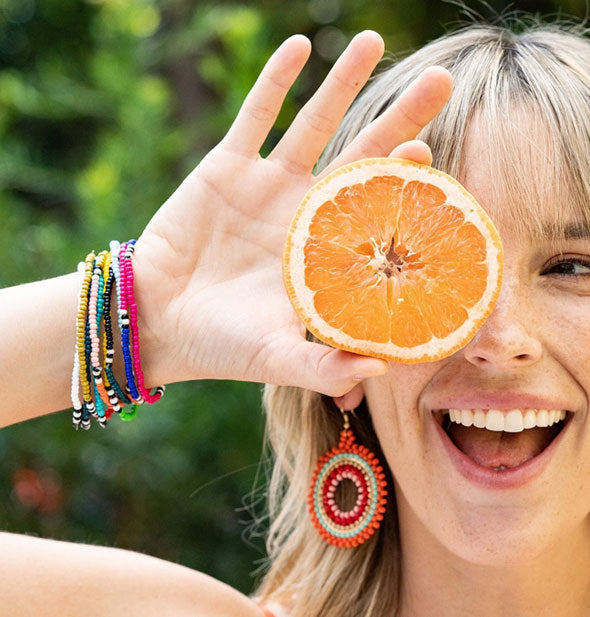 Smiling model holding half an orange over one eye wears a collection of colorful beaded bracelets on wrist