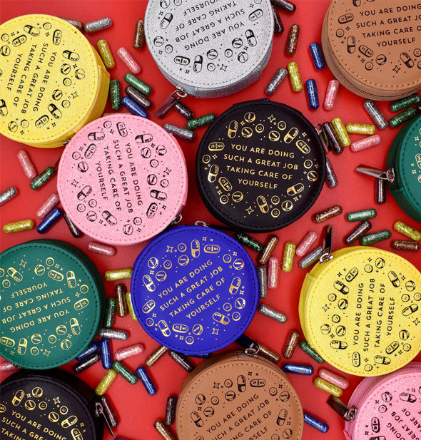 Assortment of round pill cases in a variety of colors with gold and black stamped designs around a central phrase: "You are doing such a great job taking care of yourself"; surrounding them are colorful pill capsules, all on a red surface