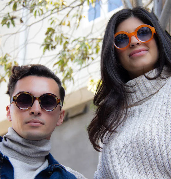 Models each wear a pair of round sunglasses, one brown tortoise with dark purplish lenses and the other orange with blue lenses