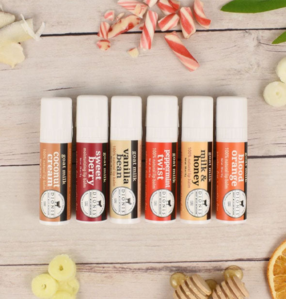 Six tubes of Dionis Goat Milk Lip Balm (Coconut Cream, Sweet Berry, Vanilla Bean, Peppermint Twist, Milk & Honey, and Blood Orange) on a distressed wooden surface scattered with fruit and candy pieces