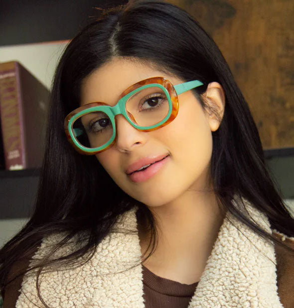 Model wears a pair of large rounded reading glasses with a two-tone green and amber tortoise frame