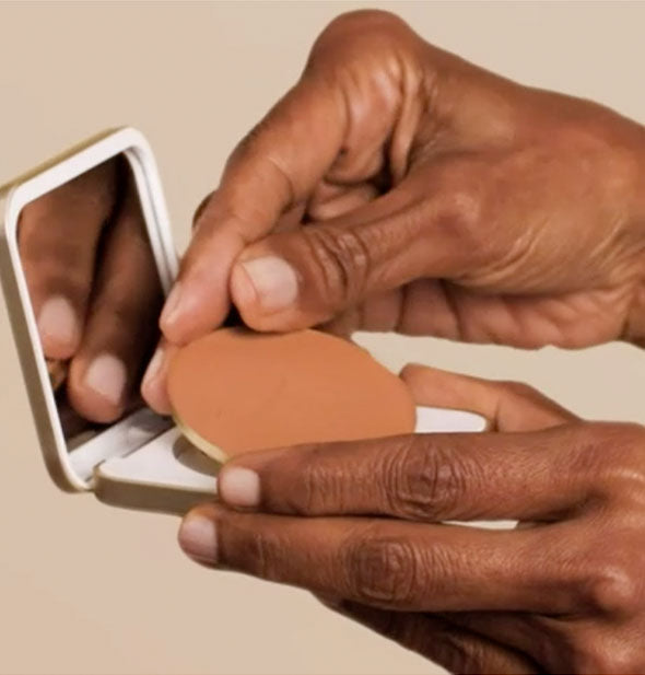 Model's hand places a round pressed powder insert in a refillable square compact with mirrored lid interior
