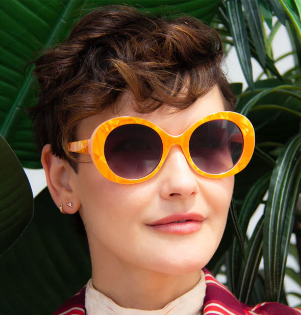 Model wears a round pair of orange sunglasses with a dark gradient lens against a backdrop of tropical plants