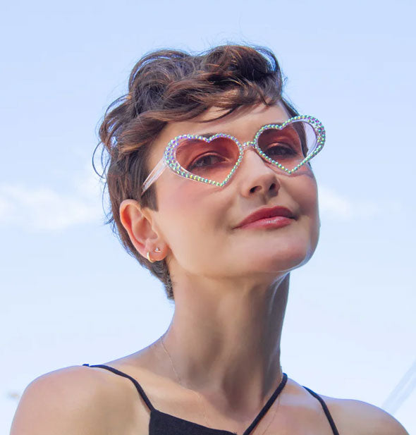 Model wears a pair of white rhinestone-encrusted diamond-shaped sunglasses with rosy-colored lenses