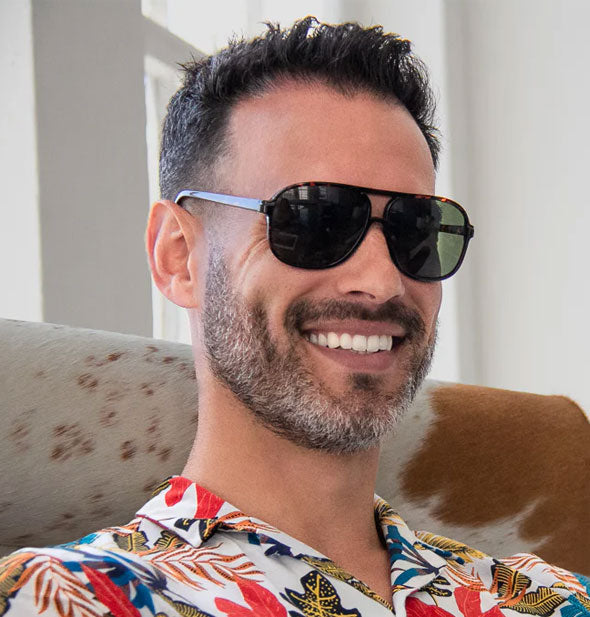 Smiling model in a tropical print shirt wears a pair of dark aviator sunglasses