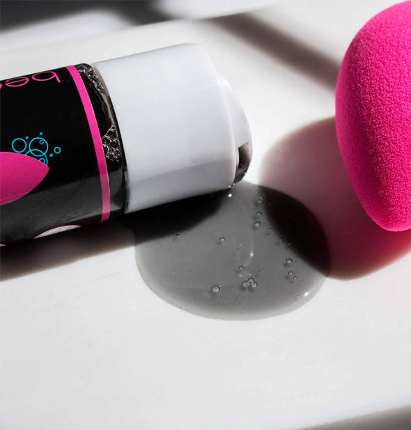 A bottle of charcoal cleanser on its side spills out grayish product onto a white surface next to a pink makeup sponge