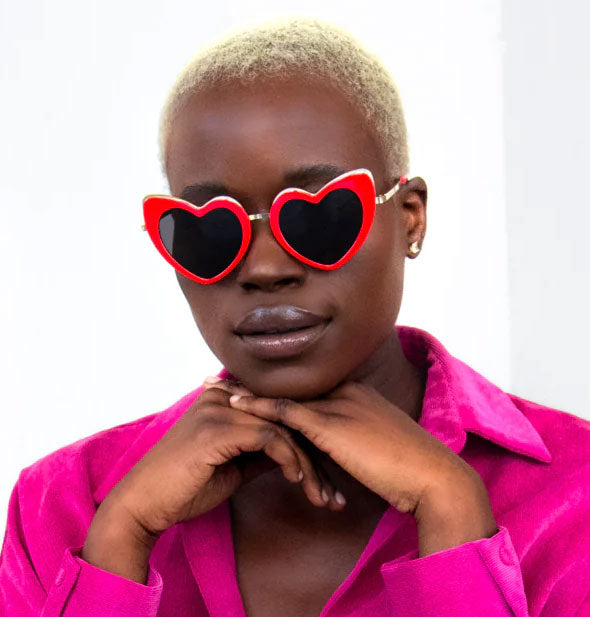 Model with hands propped under chin wears a pair of red and gold heart-shaped sunglasses with dark lenses