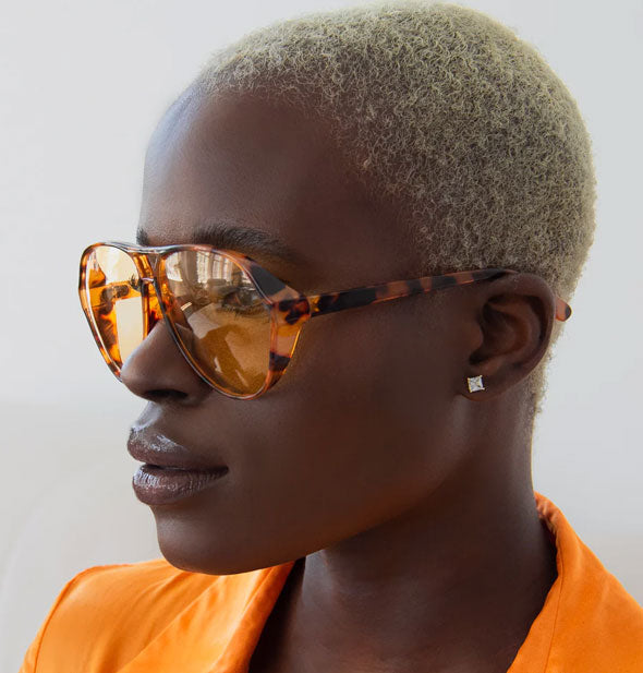 Model in orange jacket wears a pair of amber tortoise aviator sunglasses with amber lenses