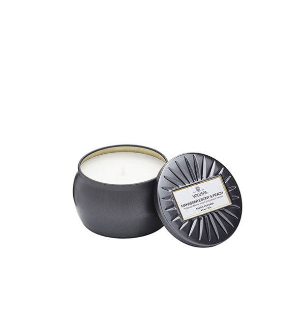 Metallic black Makassar Ebony & Peach Voluspa tin candle with lid featuring radial embossing set to the side