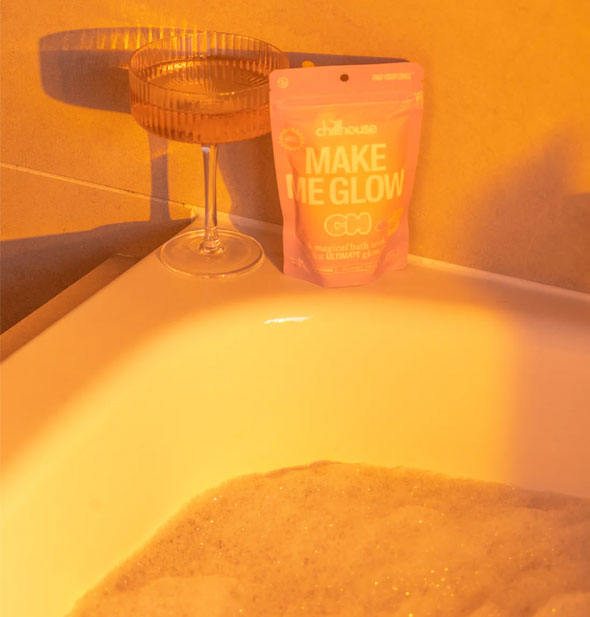 A bag of Chillhouse Make Me Glow soak rests on the edge of a tub filled with sudsy water next to a cocktail glass, all cast in an ethereal orange glow