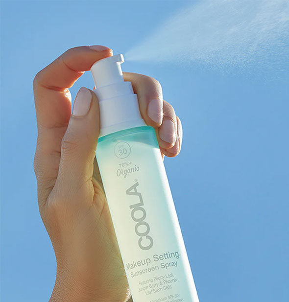 Model's hand dispenses a fine mist of Coola Makeup Setting Sunscreen Spray against a sunlit sky backdrop