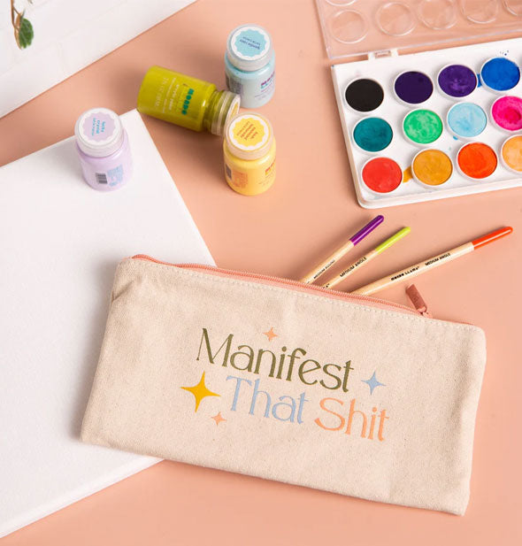 Manifest That Shit pouch on a tabletop with painting supplies