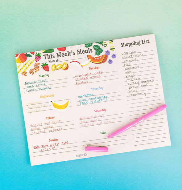 This Week's Meals and Shopping list pad with pink pen rests on a blue-green surface