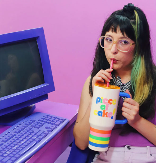 Model sitting at a pink desk with purple computer on it sips from the straw of a Piece of Cake Mega Stainless Steel Tumbler