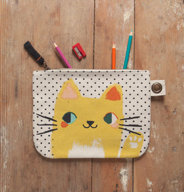 Yellow cat polkadot pouch lays on a rustic wooden surface with colored pencils sticking halfway out of it and a red pencil sharpener nearby