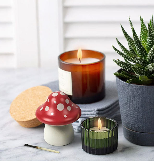 Red and white spotted ceramic mushroom match striker figurine is staged with lit candles and a potted succulent
