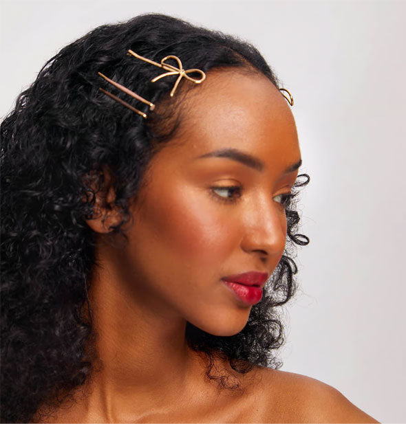 Model wears a gold bow bobby pin and two enamel covered bobby pins at the side of hair near temple