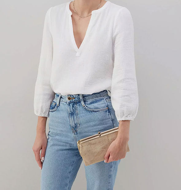 Model wearing jeans and a white shirt holds a metallic gold leather wallet with gold frame hardware for size reference