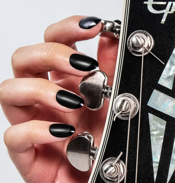 Model touches the tuning keys of a guitar with fingertips wearing a black French manicure featuring a matte base and shiny tip