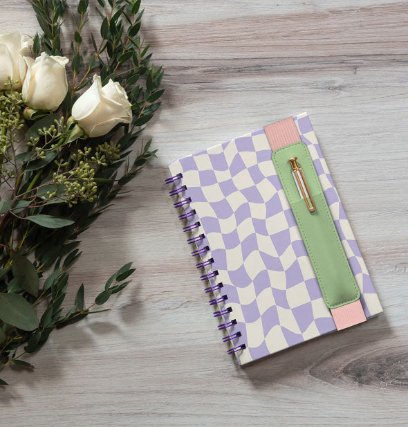 Purple and white wavy checker print spiral-bound journal with pen pocket band rests on a wooden surface next to a bouquet of white roses