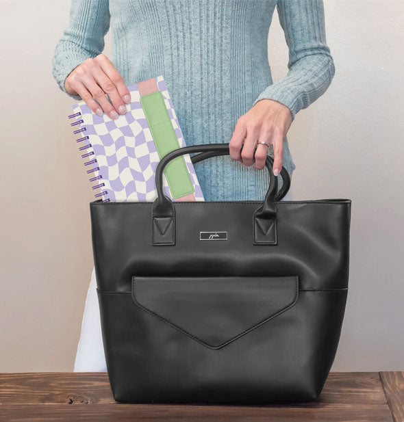 Model removes a purple and white wavy checker print notebook with pen pocket band from a black handbag