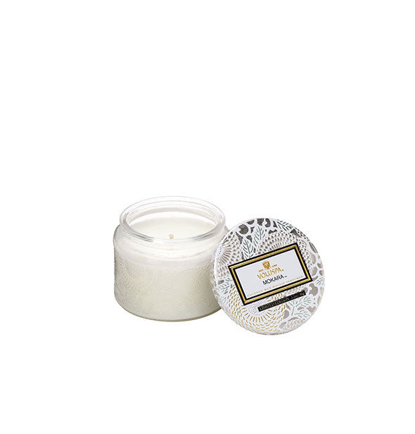 White embossed glass Mokara Volspa jar candle with white, silver, and gold metallic floral lid set to the side