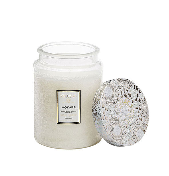 White embossed glass Mokara Voluspa candle jar with white and gold metallic floral lid set to the side