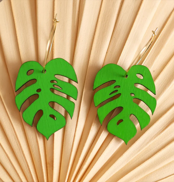 A pair of green monstera leaves on gold earring hoops rest on a bamboo fan