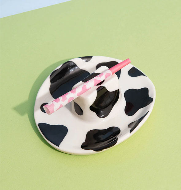 Black and white cowhide print cowboy hat ashtray rests on a green surface and holds a pink rolled cigarette in its top groove