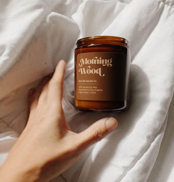 Model's hand reaches for a Morning Wood amber glass jar candle resting on a bedspread