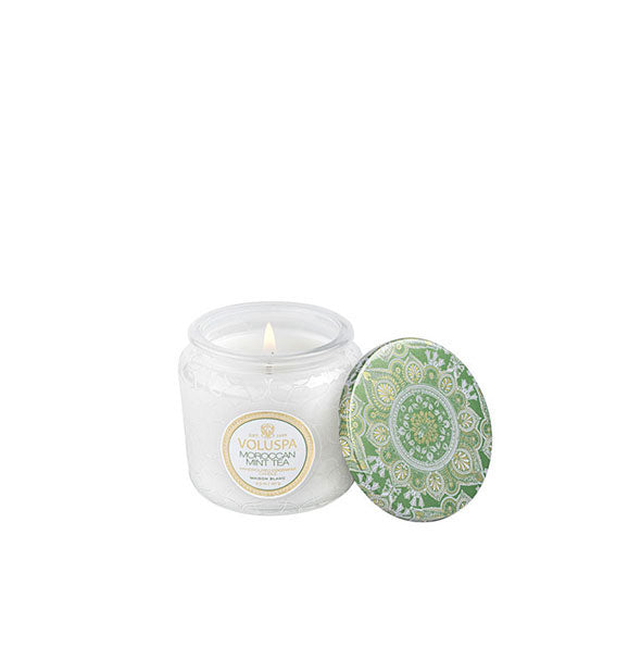 A lit Voluspa Moroccan Mint Tea candle in frosted embossed glass jar with decorative green tin lid alongside