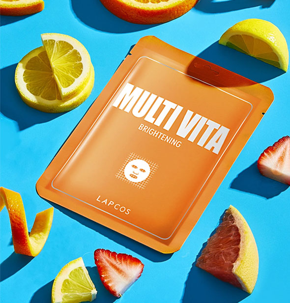 Orange Multi Vita Brightening mask packet rests on a blue surface surrounded by pieces of fruit
