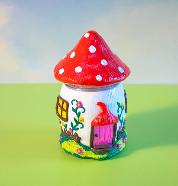 A colorful jar designed to resemble a mushroom house with red and white spotted "cap" lid, pink door, and flower vine details