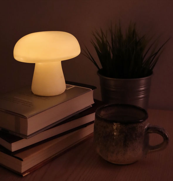 Lit-from-within white mushroom-shaped lamp rests on a small stack of books next to a coffee mug and potted plant in a darkened room