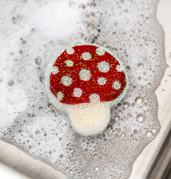 Red and white speckled mushroom dish sponge resting in soap suds