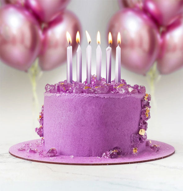 Six purpleish crystal-shaped lit candles decorate a purple cake with metallic balloons in the background