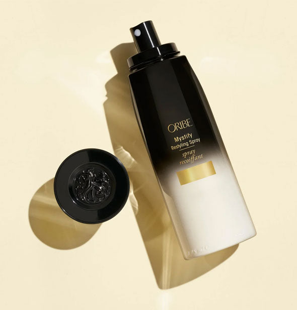 A bottle of Oribe Mystify Restyling Spray lays on its side with cap removed and placed neatby