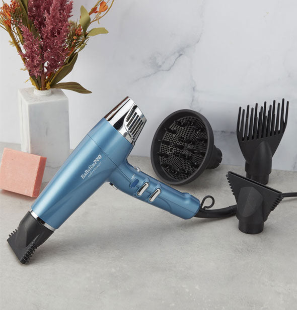 Blue and silver BaBylissPRO Nano Titanium hair dryer rests on a countertop with its included black attachments: 2 concentrator nozzles (one of which has been placed on the end of the dryer's nozzle), pronged straightening pick, and round diffuser