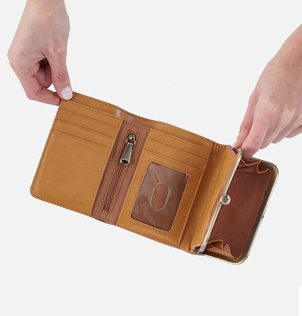 Model's hands hold open a honey brown leather tri-fold wallet with antique brass frame hardware and ample interior storage space