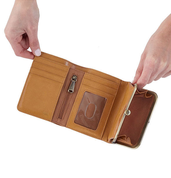 Model's hands hold open a honey brown leather tri-fold wallet with antique brass frame hardware and ample interior storage space