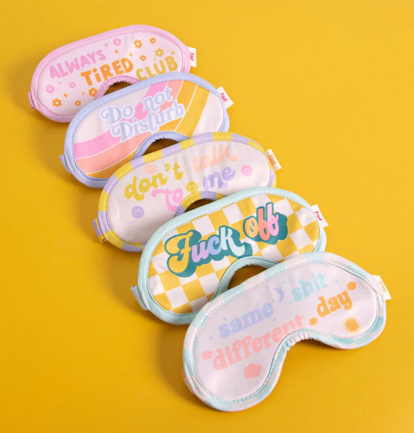 Collection of five sleep masks in assorted pastel designs with sassy messages rest on a bright yellow backdrop