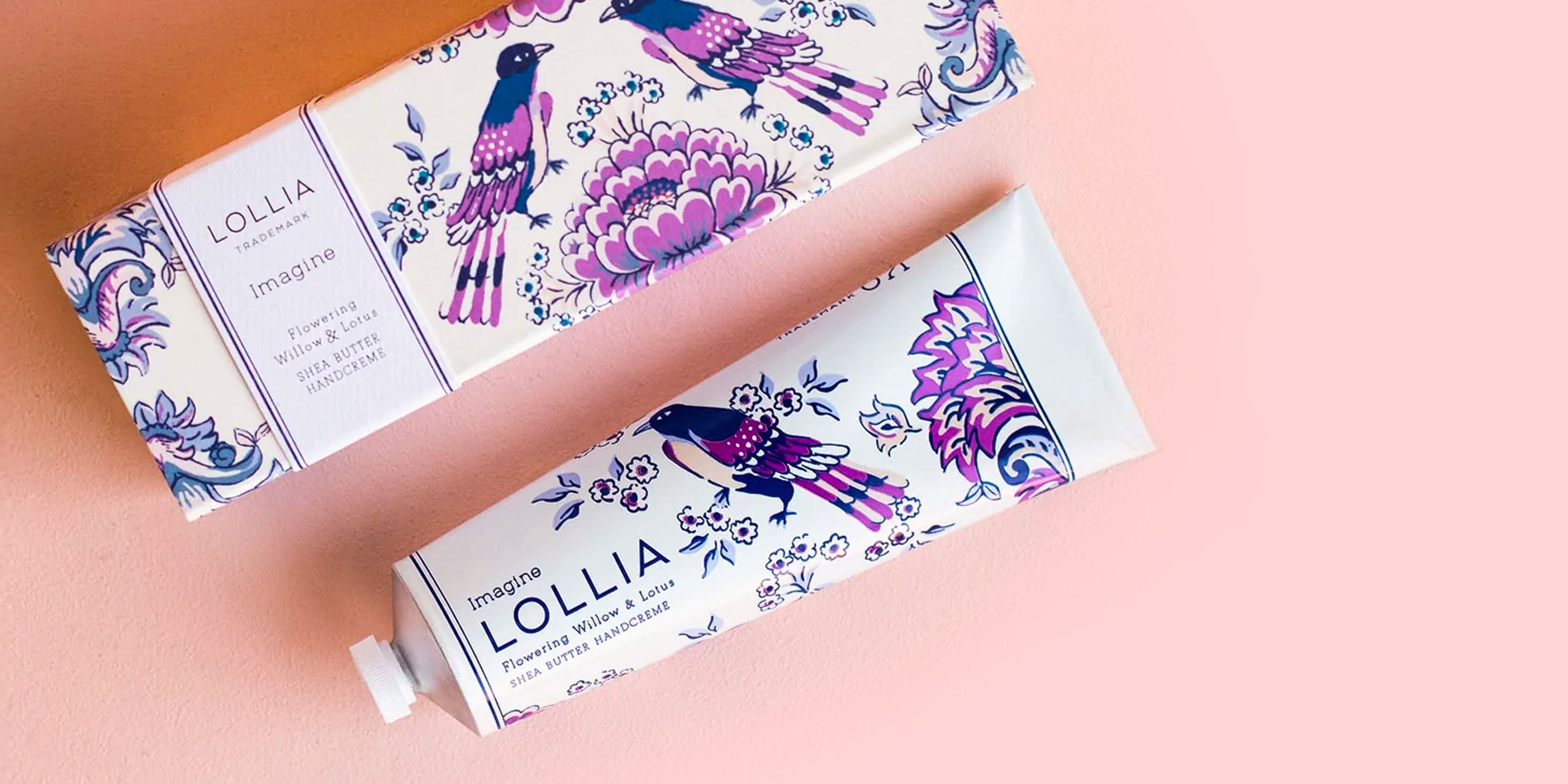 A tube and box of Lolla hand cream printed with decorative purple birds and florals rest on a pink surface